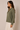 A woman wearing a muted olive green long-sleeve button-up shirt and white pants, shown in profile against a plain beige studio background.