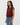 A woman wearing a sleeveless, gathered maroon top and blue jeans, photographed from the waist up against a plain light gray studio background.