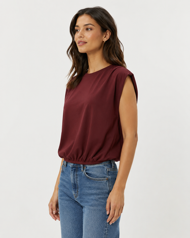 A woman wearing a sleeveless, gathered maroon top and blue jeans, photographed from the waist up against a plain light gray studio background.