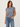 A woman wearing a muted slate blue-purple short-sleeve top with gathered shoulder details and blue jeans, standing against a plain light gray studio background.