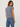 A woman shown from the back wearing a sleeveless dusty blue top and blue jeans against a plain light gray studio background.