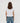 A woman viewed from behind wearing a white short-sleeve top and blue jeans against a plain light gray studio background.
