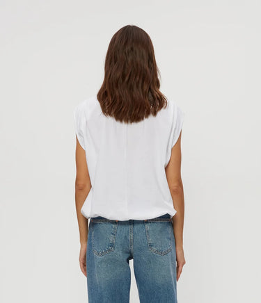 A woman viewed from behind wearing a white short-sleeve top and blue jeans against a plain light gray studio background.