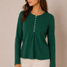 A woman wearing a dark green long-sleeve ribbed knit top with a buttoned neckline and cream pants, standing against a plain beige studio background.