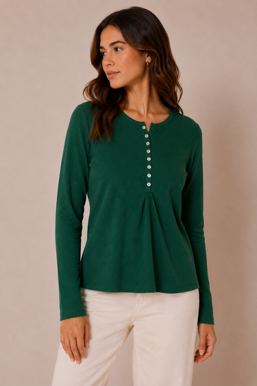 A woman wearing a dark green long-sleeve ribbed henley top with a button-down front and white pants, standing against a plain beige studio background.