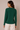 A woman shown from the back wearing a dark green ribbed long-sleeve top and cream-colored pants, standing against a solid beige studio background.