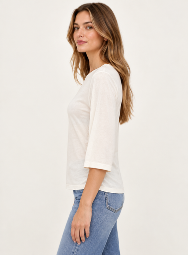 A woman wearing a white three-quarter sleeve t-shirt and blue jeans, shown from a side profile against a plain off-white studio background.
