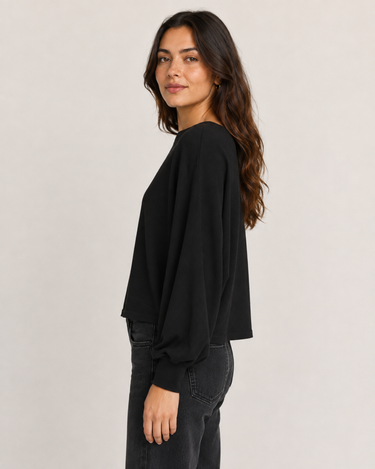 A woman wearing a black long-sleeve top with a relaxed, draped fit and dark gray jeans, photographed against a plain light gray studio background.