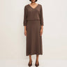 A woman modeling a solid brown midi dress with a V-neckline, three-quarter sleeves, and a draped overlay, paired with brown pointed-toe heels against a plain off-white studio background.