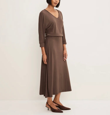 A woman modeling a two-piece brown dress set consisting of a long-sleeve V-neck top and a matching midi skirt, paired with brown pointed-toe heels against a plain off-white studio background.