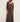 A woman wearing a long, draped brown maxi dress with three-quarter sleeves and a V-neckline, photographed against a plain off-white wall.