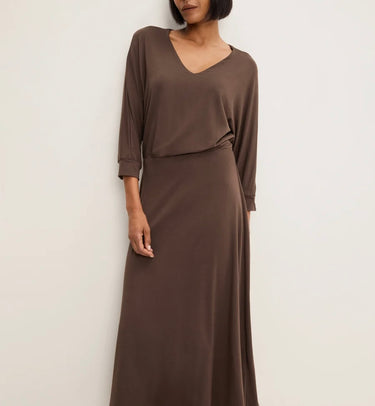 A woman wearing a long, draped brown maxi dress with three-quarter sleeves and a V-neckline, photographed against a plain off-white wall.