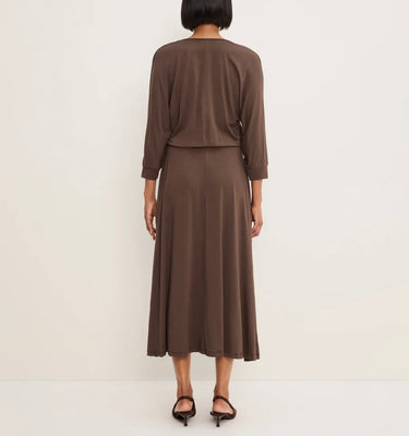 A woman viewed from the back wearing a brown long-sleeve top and matching midi skirt set, paired with black heeled sandals, against a plain off-white studio background.