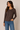 A woman wearing a dark brown long-sleeve button-front top and blue jeans, standing against a plain beige studio background.