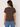 A woman viewed from the back wearing a dark brown short-sleeve crewneck t-shirt and blue jeans, standing against a plain off-white studio background.