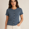 A woman modeling a short-sleeve crewneck top in heathered slate blue, featuring a button-down front with cream buttons. She is wearing off-white pants against a plain beige studio background.