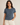 A woman modeling a short-sleeve crewneck top in heathered slate blue, featuring a button-down front with cream buttons. She is wearing off-white pants against a plain beige studio background.
