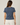 A woman viewed from the back wearing a heathered slate blue short-sleeve t-shirt and off-white pants, set against a plain light beige studio background.