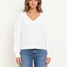 A woman facing foreward wearing a white v-neck long sleeve and blue jeans on a white background.