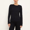 A woman wearing a black long-sleeve crewneck top and black pants, standing against a plain off-white background.