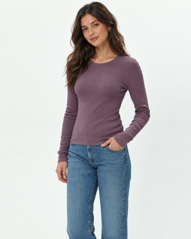 A woman modeling a long-sleeve crewneck knit top in a dusty mauve-purple color, paired with blue jeans, against a clean white studio background.