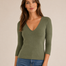 A woman modeling a fitted olive green long-sleeve V-neck top with three-quarter sleeves, paired with blue jeans, against a plain beige studio background.