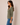 A woman wearing a sage green three-quarter sleeve V-neck top and blue jeans, shown in profile against a plain beige studio background.