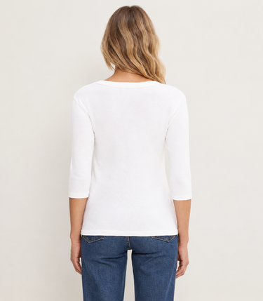 A woman viewed from the back wearing a white three-quarter sleeve top and blue jeans, standing against a plain off-white studio background.