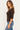 A woman wearing a brown ribbed 3/4-sleeve scoop-neck top and blue jeans, standing against a plain off-white background.