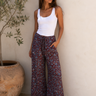 Woman wearing a white tank top and patterned pants standing against a neutral wall with a plant in the background.