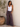 Woman wearing a white tank top and patterned pants standing against a neutral wall with a plant in the background.