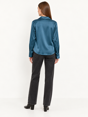 A woman viewed from the back wearing a teal satin long-sleeve blouse, dark gray jeans, and black ankle boots against a plain white studio background.
