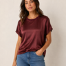 A woman wearing a short-sleeve crewneck satin top in a deep burgundy color, paired with blue jeans, standing against a plain light beige background.