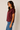 A woman wearing a short-sleeve satin top in burgundy and blue jeans, standing against a plain light beige studio background.