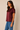 A woman wearing a short-sleeve satin top in deep burgundy with blue jeans, standing against a plain beige studio background.
