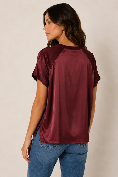 A woman shown from the back wearing a short-sleeve satin-like top in deep burgundy with contrasting darker burgundy raglan sleeves, paired with blue jeans, against a plain light beige studio background.