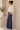 Woman wearing a white blouse and blue pants standing in a minimalistic room with a vase of plants.