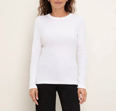 A woman wearing a fitted white long-sleeve crewneck top and black pants, standing against a plain light beige background.