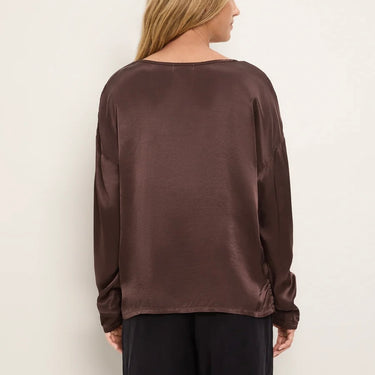 A woman viewed from the back wearing a dark brown long-sleeve satin blouse and black pants, set against a plain off-white background.
