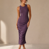 A woman modeling a sleeveless, ruched midi dress in a muted purple color, paired with clear high-heeled sandals, set against a neutral beige studio backdrop.