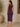 A woman wearing a sleeveless, knee-length purple dress, shown from the back in a warm indoor setting with a potted plant and wooden wall decor.