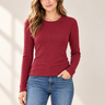 A woman modeling a long-sleeve crewneck knit top in deep burgundy, paired with blue jeans, against a warm beige background with soft window light shadows.