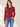 A woman modeling a long-sleeve crewneck knit top in deep burgundy, paired with blue jeans, against a warm beige background with soft window light shadows.