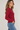 A woman wearing a long-sleeve crewneck sweater in deep burgundy, paired with blue jeans, shown from a side profile against a light gray studio background.