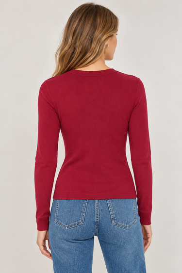 A woman viewed from the back wearing a fitted long-sleeve crewneck top in deep red and blue jeans, against a plain light gray studio background.
