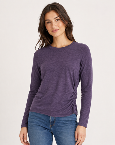 A woman modeling a heathered purple long-sleeve top with a gathered side detail, paired with blue jeans, against a plain light background.