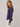A woman wearing a purple long-sleeve top with ruching on the side and white pants, photographed against a light gray textured studio background.