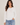 A woman modeling a white long-sleeve crewneck top with side ruching, paired with light blue jeans, against a plain light beige studio background.