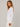 A woman wearing a white long-sleeve top with a ruched side detail and white pants, photographed against a plain light gray studio background.