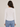 A woman viewed from the back wearing a white long-sleeve crewneck top and light blue jeans, set against a plain light beige studio background.